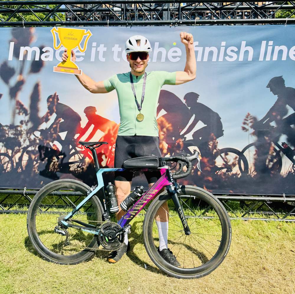 Sven holding a trophy and raising a fist in front of an 'I made it to the finish line' banner, road bike in front
